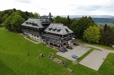 Zdjęcie wykonane z dużej wysokości. Na zdjęciu kilkukondygnacyjny budynek ze spadzistym dachem.  W tle ściana wysokich drzew, a w oddaleniu zarys gór.