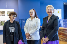 Trzy kobiety uśmiechają się do zdjęcia. W tle ciemno niebieska ściana i plansze ze zdjęciami.