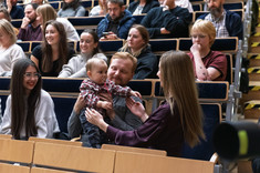 Zbliżenie na uczestników wykładu siedzących w rzędach. Pośrodku mała dziewczynka, najmłodszy uczestnik wykładu. Wszyscy się uśmiechają.