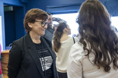 Na pierwszym planie dwie kobiety, jedna ubrana na biało z długi ciemnymi włosami, odwrócona plecami, druga z krótkimi włosami ubrana na czarno. 