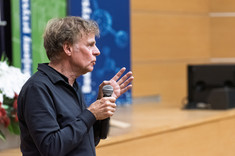 Mężczyzna ubrany na czarno podczas prowadzenia wykładu. W tle zielono-biało-niebieskie banery i drewnopodobna ściana sali wykładowej.