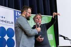 Dwóch mężczyzn prowadzi wykład. Starszy mężczyzna ubrany na czarno tłumaczy coś zebranym, drugi w jasnej marynarce stoi obok. 