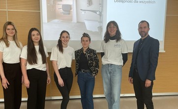 Na tle ekranu z wyświetloną prezentacją grupa sześciu osób. Po lewej mężczyzna w ciemnym ubraniu. Pośrodku grupy kobiet w białych podkoszulkach prelegentka w ciemnym ubraniu. Wszyscy się uśmiechają.