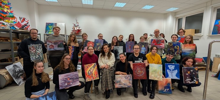 W jasno oświetlonej sali około 30 osób pozujących do wspólnego zdjęcia. Każdy trzyma w rękach barwny obraz.