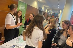 Na zdjęciu widać grupę młodych osób przy stoisku promocyjnym. Dwie osoby w białych koszulkach rozmawiają z zainteresowanymi. W tle baner „Katedra Urbanistyki i Architektury”. Korytarz jest jasny, pełen studentów.