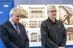 Dwóch mężczyzn na ciemno ubranych stoi na tle planszy ze zdjęciami. 