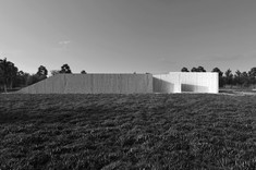 Pozioma, prostokątna plansza przedstawia czarno-białą wizualizację projektowanego budynku. Prosta betonowa bryła na tle naturalnego krajobrazu.