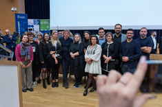 Zdjęcie grupowe kilkunastu osób, mężczyzn i kobiet. Większość ubrana na czarno. Wszyscy się uśmiechają. W tle scena sali wykładowej.