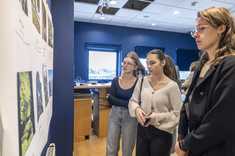 Na pierwszym planie trzy młode kobiety przyglądają się białej planszy z kolorowymi zdjęciami.