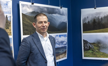Na pierwszym planie stoi autor wystawy, mężczyzna w ciemnej marynarce.  W tle na granatowej ścianie zawieszone plansze z dużymi kolorowymi fotografiami pejzaży górskich.