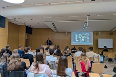 Wewnątrz sali z jasnym drewnopodobnym wykończeniem ścian i białym sufitem zgromadzonych jest kilkadziesiąt osób podczas wykładu. Na zdjęciu w oddaleniu widoczny jest ekran z prezentacją i prelegent, wszyscy słuchacze siedzą. 