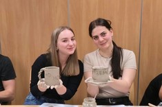 Na zdjęciu dwie młode uśmiechnięte kobiety z długimi włosami. Obie trzymają w rękach ulepione z gliny kubki. 