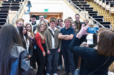 Zdjęcie grupowe studentów, około dwudziestu osób, młodych mężczyzn i kobiet. Po środku wykładowca, starszy mężczyzna ubrany na czarno. Wszyscy się uśmiechają. W tle wyjście z sali wykładowej.