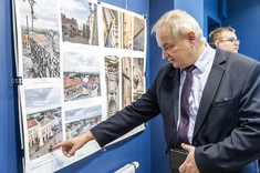 Mężczyzna w ciemnym garniturze wskazuje dłonią fragment wywieszonego zdjęcia.