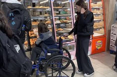 Zdjęcie przedstawia studentów podczas zajęć, którzy robią zakupy w sklepie. Jedna studentka korzysta z wózka inwalidzkiego próbując dosięgnąć artykułów znajdujących się na wyższych regałach 