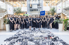 W centrum ujęcia na podłodze ułożony kwadrat około 3 x 3 metry z biało-czarnych kwadratowych makiet. Wokół zgromadzona grupa kilkudziesięciu osób. Wszyscy ubrani na czarno. 