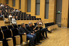 Zdjęcie ukazuje fragment audytorium podczas wydarzenia. Widzimy przestronną salę z nowoczesnym, drewnianym wystrojem ścian i dużymi, zasłoniętymi oknami.  W centralnej części kadru siedzi grupa uczestników — w pierwszym rzędzie zasiada kilka osób. Są elegancko ubrani, a część z nich jest zaangażowana w rozmowę.