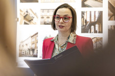 Kobieta o ciemnych włosach, w okularach, ubrana w czerwoną marynarkę, stoi na tle planszy ze zdjęciami.