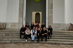 Grupa dziesięciu osób siedzi koło siebie na szerokich schodach wejściowych do budynku.  W tle wejście do budynku, jasna wysoka ściana z wysokim zakończonym łukiem wejściem i wyraźnym napisem POLONIA.