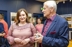 Na pierwszym planie w pomieszczeniu stoją dwie osoby, kobieta w różowej bluzce i mężczyzna w ciemnej marynarce. Rozmawiają ze sobą. W tle stoi kilka osób.