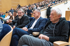 Duża grupa osób na sali wykładowej. Na pierwszym planie siedzi koło siebie trzech mężczyzn.