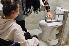 Zdjęcie przedstawia dwie osoby w toalecie. Jedna na wózku inwalidzkim, druga w ortezach na nogach i rękach, próbująca skorzystać z toalety. 