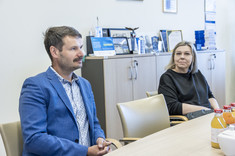 Kobieta i mężczyzna siedzą przy stole. Kobieta delikatnie uśmiecha się. W tle jasna ściana.