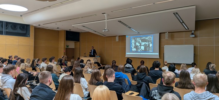 Wewnątrz sali z jasnym drewnopodobnym wykończeniem ścian i białym sufitem zgromadzonych jest kilkadziesiąt osób podczas wykładu. Na zdjęciu w oddaleniu widoczny jest ekran z prezentacją i prelegent, wszyscy słuchacze siedzą. 